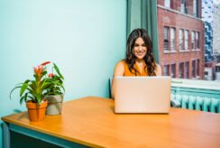 Confident woman in a bright office working on a laptop, surrounded by urban scenery and greenery.